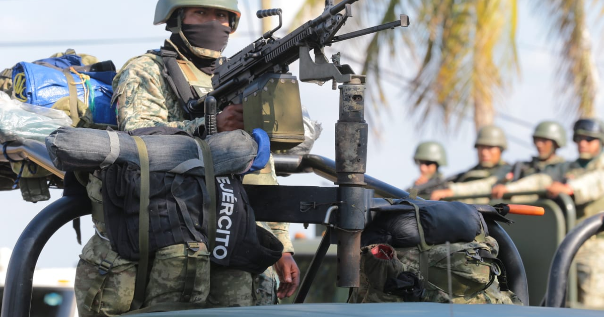 Soldado armado con mochila y equipo sobre vehículo militar en entorno tropical, con soldados en formación al fondo.