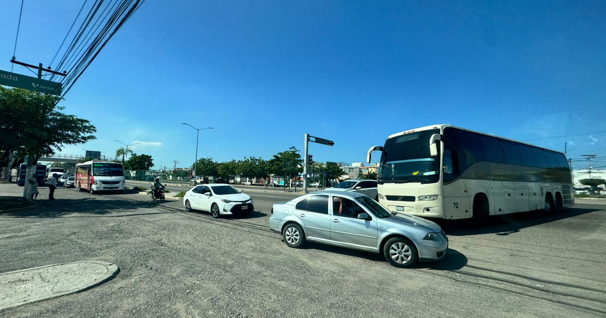 Intersección con tráfico vehicular en carretera internacional, incluyendo autobús, coches y motocicleta, con cielo despejado.