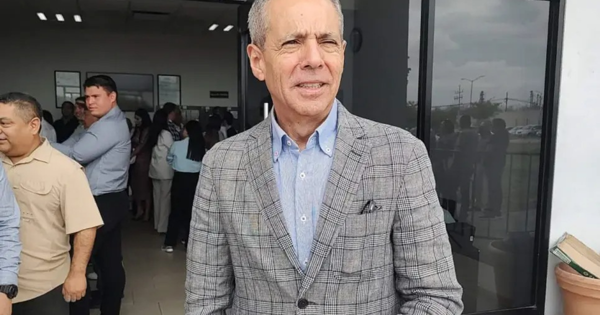 Hombre con saco de cuadros en evento oficial, rodeado de personas en oficina iluminada.