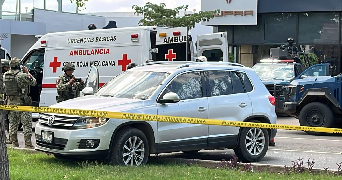 SUV plateado con vidrios rotos tras ataque armado en Culiacán, rodeado de seguridad y ambulancia.