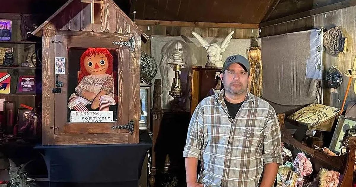 Hombre junto a caja de madera con muñeca Annabelle, rodeado de objetos paranormales.