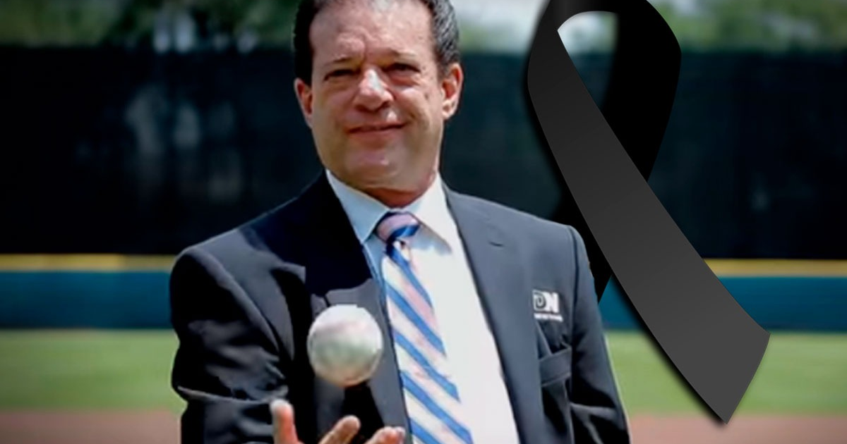 Hombre con traje y corbata sosteniendo una pelota en un campo de béisbol, con un lazo negro en la esquina.