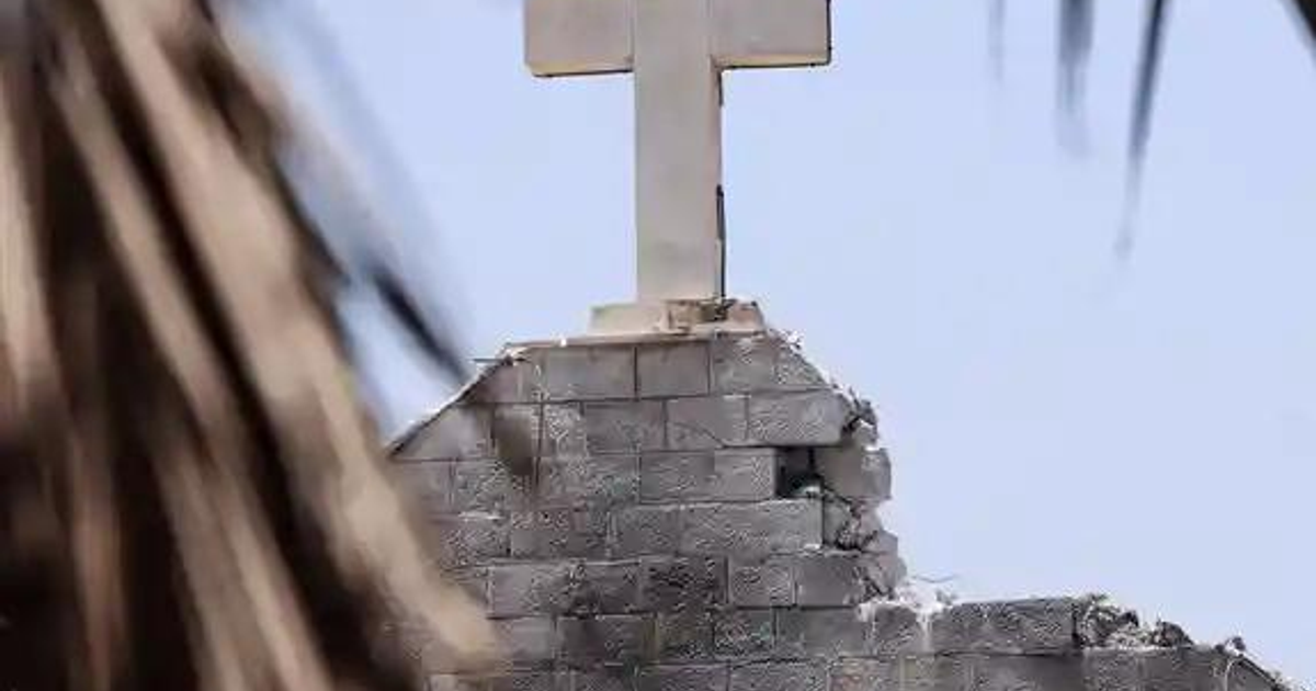 Cruz blanca sobre estructura en ruinas de la iglesia de la Sagrada Familia en Gaza, con cielo azul y hojas en primer plano.