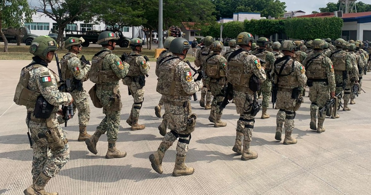 Soldados marchando en una base militar con uniformes camuflados y vehículos en el fondo.