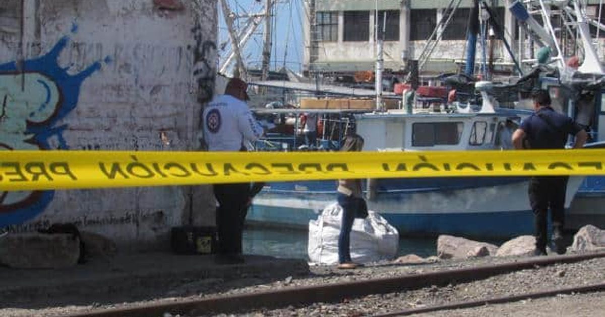Área de puerto con barco, personas y cinta de precaución, sugiriendo investigación o emergencia.