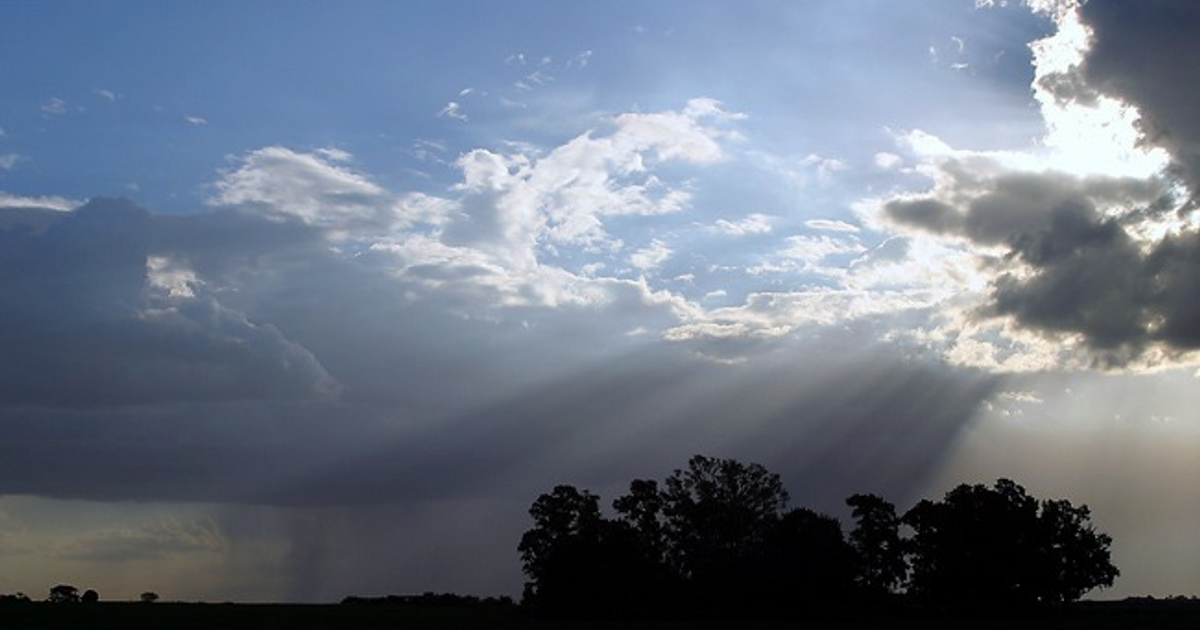 Cielo nublado con rayos de luz filtrándose, tonos azules y grises, sombras de árboles en la parte inferior.
