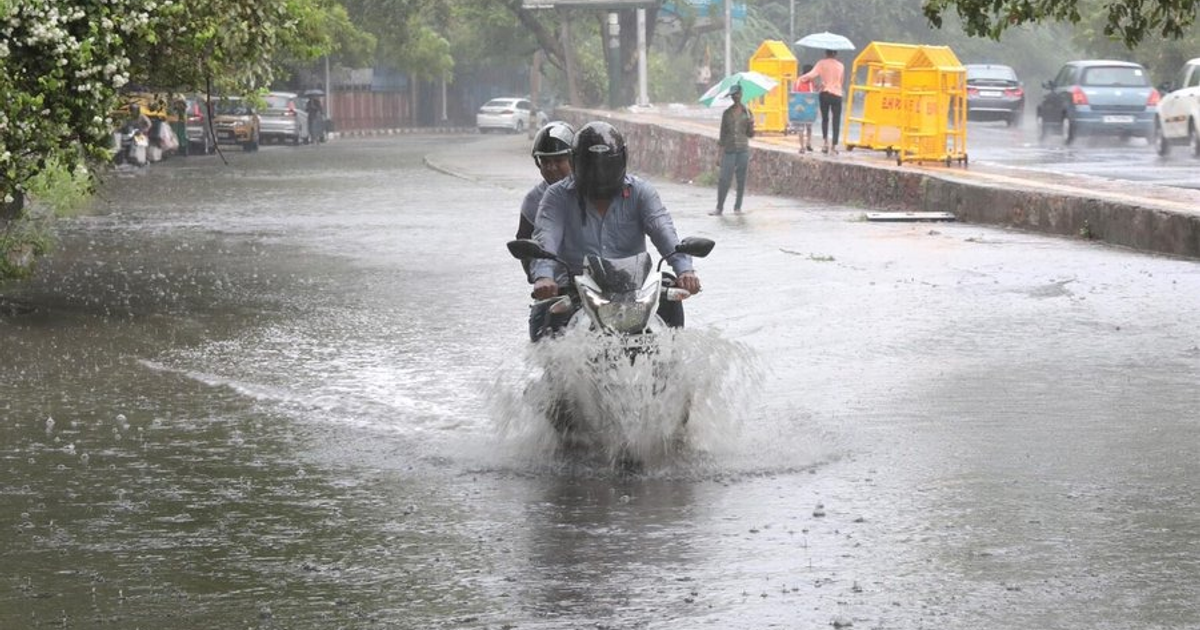 Dos personas en motocicleta cruzan una calle inundada bajo la lluvia, con charcos y salpicaduras alrededor, en un paisaje urbano lluvioso.