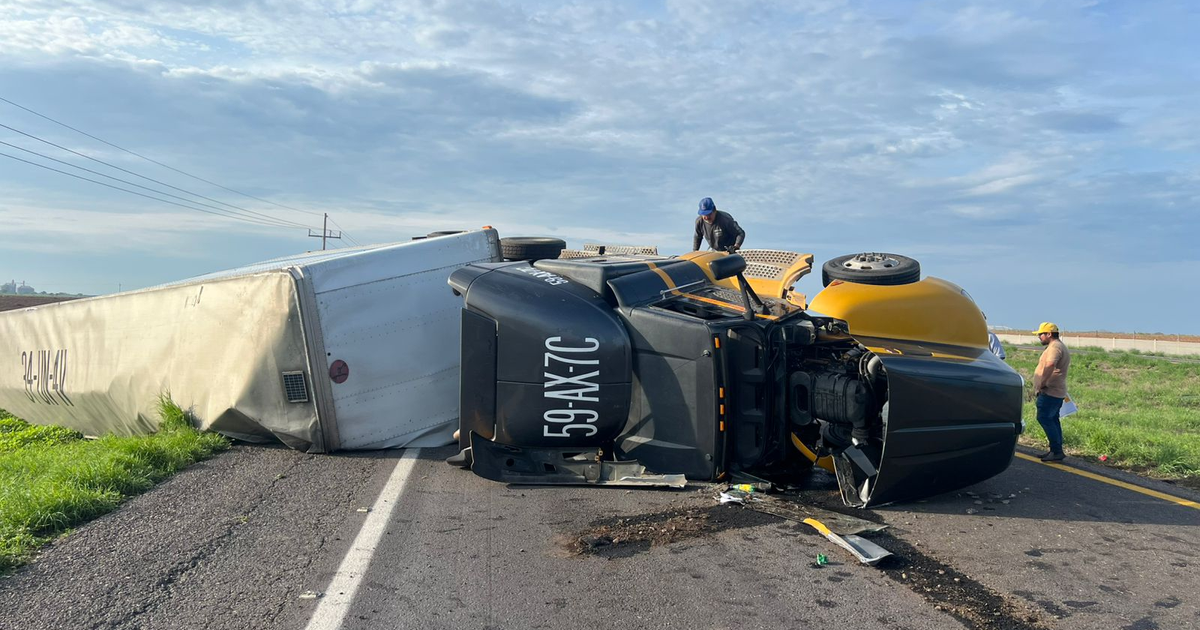 Camión volcado en carretera con dos personas inspeccionando, cielo nublado y hierba alrededor.