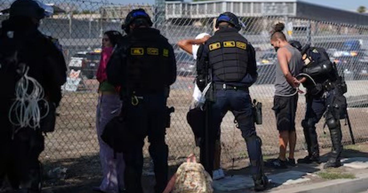 Agentes de seguridad detienen a migrantes en Florida, con una cerca visible al fondo, en un operativo policial relacionado con el aumento de detenciones tras el regreso de Trump a la presidencia.