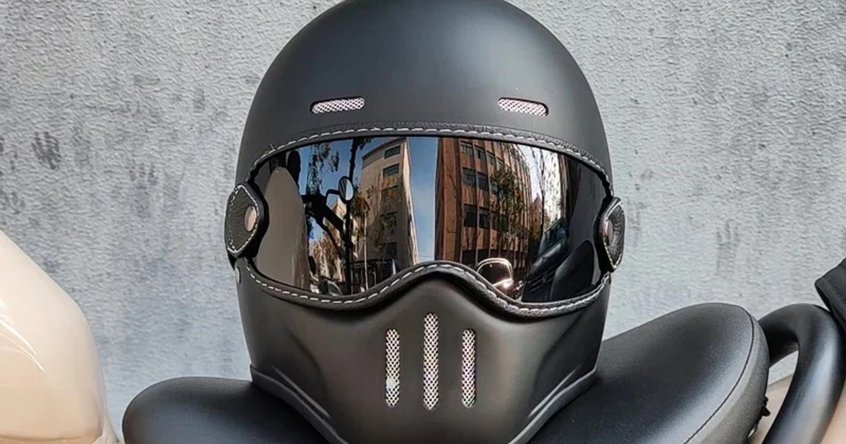 Casco de motocicleta negro mate con visera reflectante sobre asiento de moto, edificios reflejados en la visera.