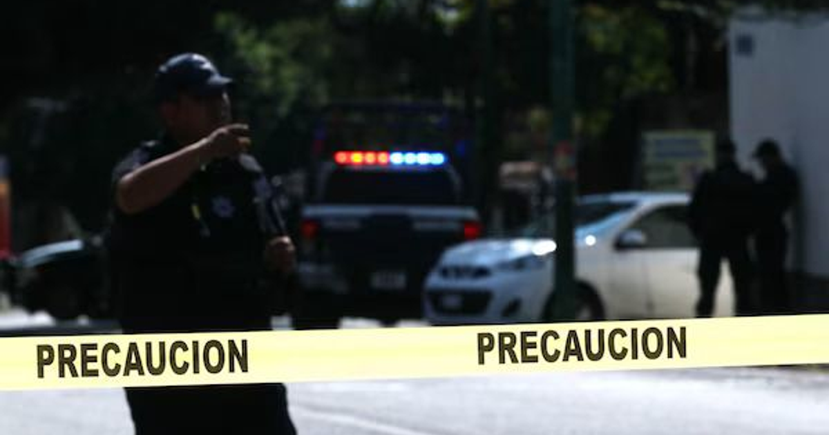 Oficial de policía en escena de investigación urbana con cinta de precaución y vehículo policial al fondo.