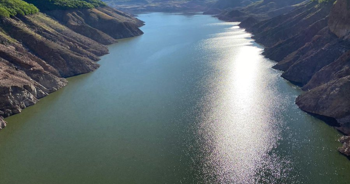 Embalse en Sinaloa con reflejos solares y montañas alrededor, mostrando un paisaje árido con vegetación verde.