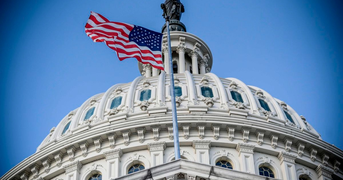 Cúpula del Capitolio de EE.UU. con bandera estadounidense ondeando, cielo azul claro de fondo.