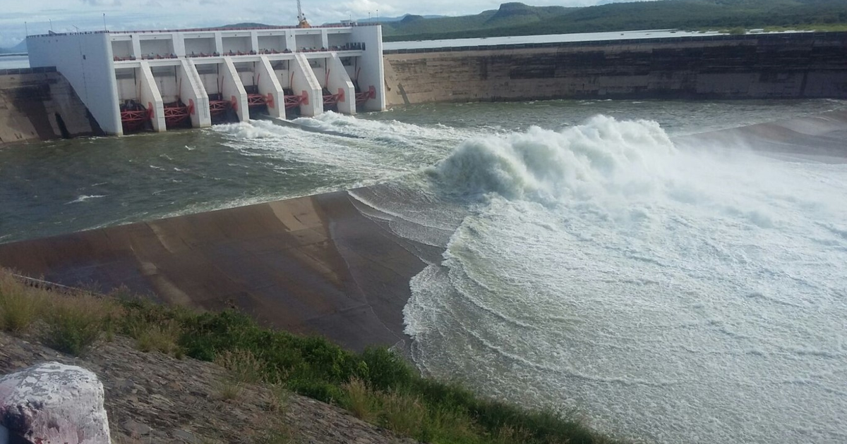 Represa en Sinaloa con agua fluyendo rápidamente desde compuertas, cielo parcialmente nublado y vegetación circundante.