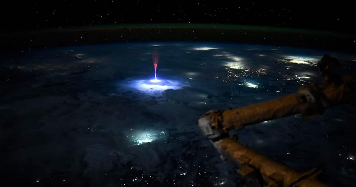 Vista espacial de un sprite rojo sobre la Tierra, capturado desde la Estación Espacial Internacional, con un brazo robótico en primer plano y un fondo estrellado.