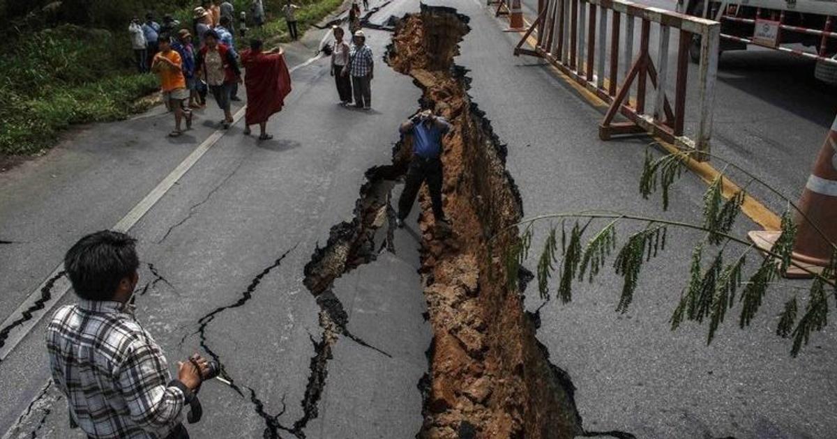 Carretera con deslizamiento de tierra y grieta profunda, personas observando y tomando fotos.