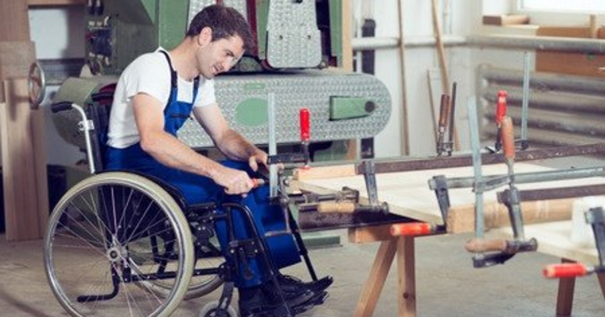 Persona en silla de ruedas trabajando en carpintería con herramientas y maquinaria de fondo.