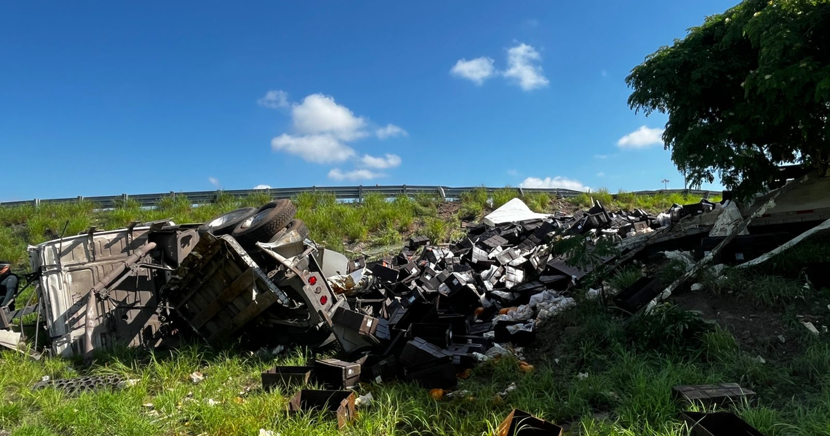 Camión volcado en terreno junto a carretera con cajas esparcidas, cielo azul y nubes.