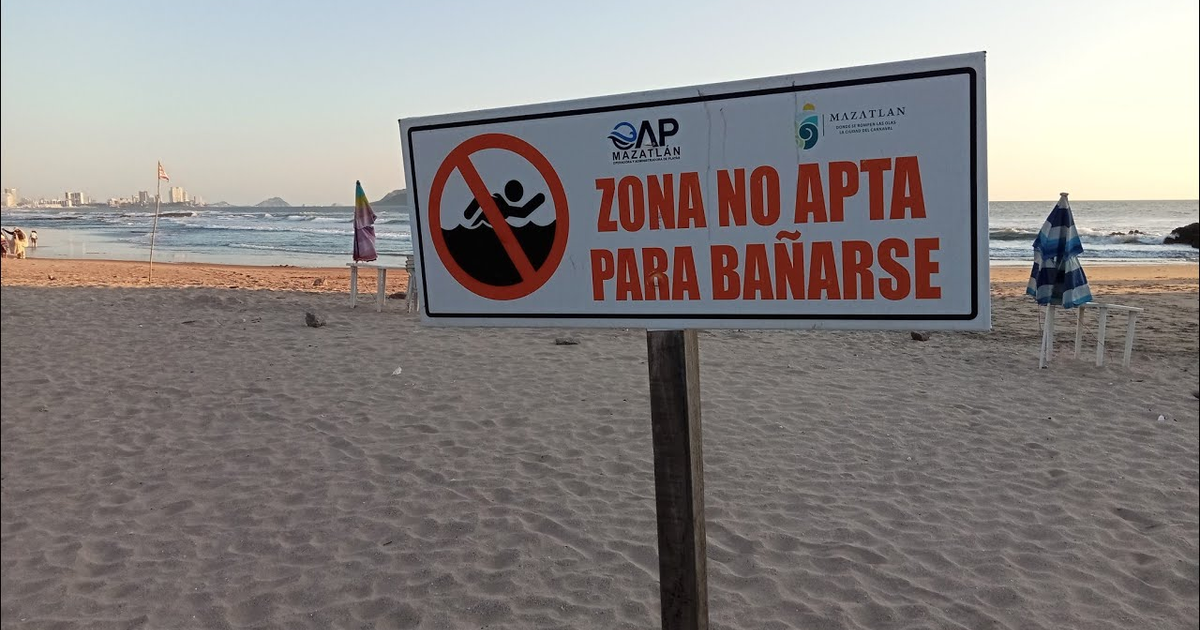 Cartel de advertencia en playa de Mazatlán, zona no apta para bañarse, con sombrillas y mar al fondo.