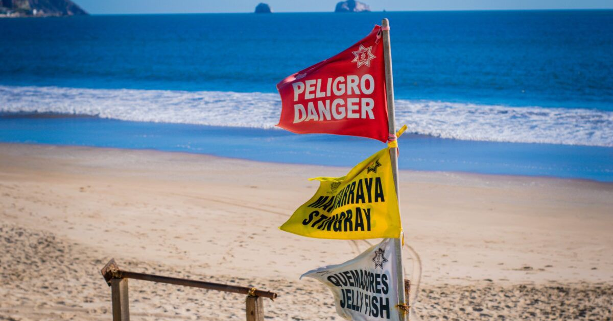 Banderas de advertencia en playa de Mazatlán con indicaciones de peligro, mantarrayas y medusas.