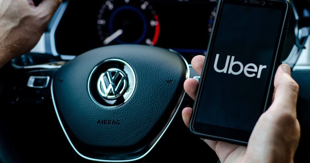 Mano sosteniendo un teléfono con la app de Uber frente a un volante de Volkswagen, en el contexto del debate sobre costos laborales en plataformas digitales.
