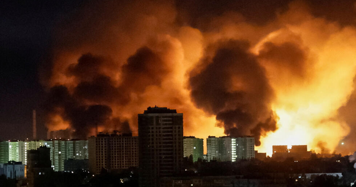 Paisaje nocturno con incendio, humo negro y edificios iluminados.