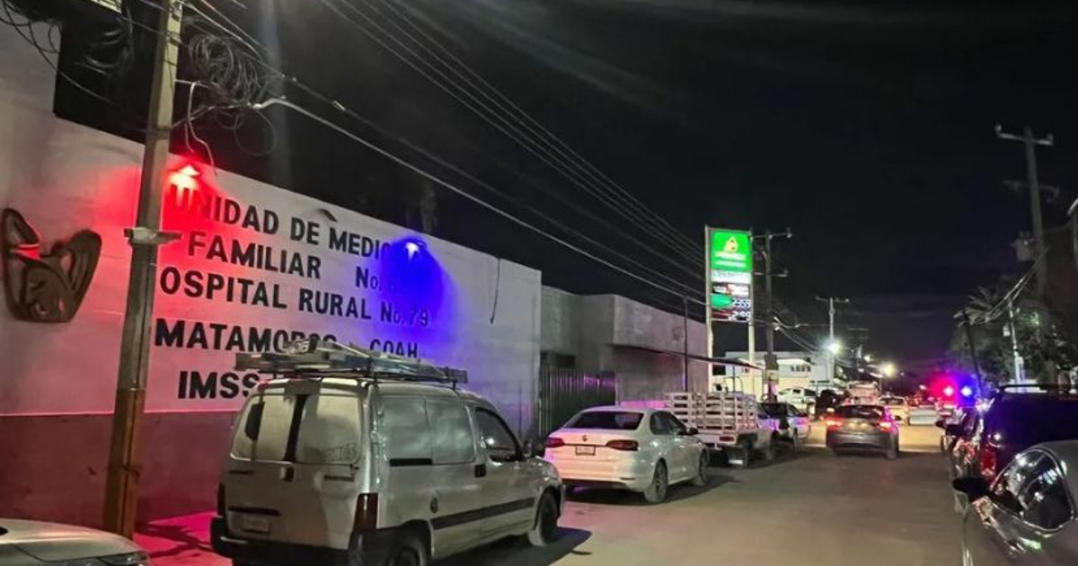 Calle nocturna con hospital rural y unidad de medicina familiar iluminada por luces de colores, con vehículos estacionados y letrero de gasolinera al fondo.