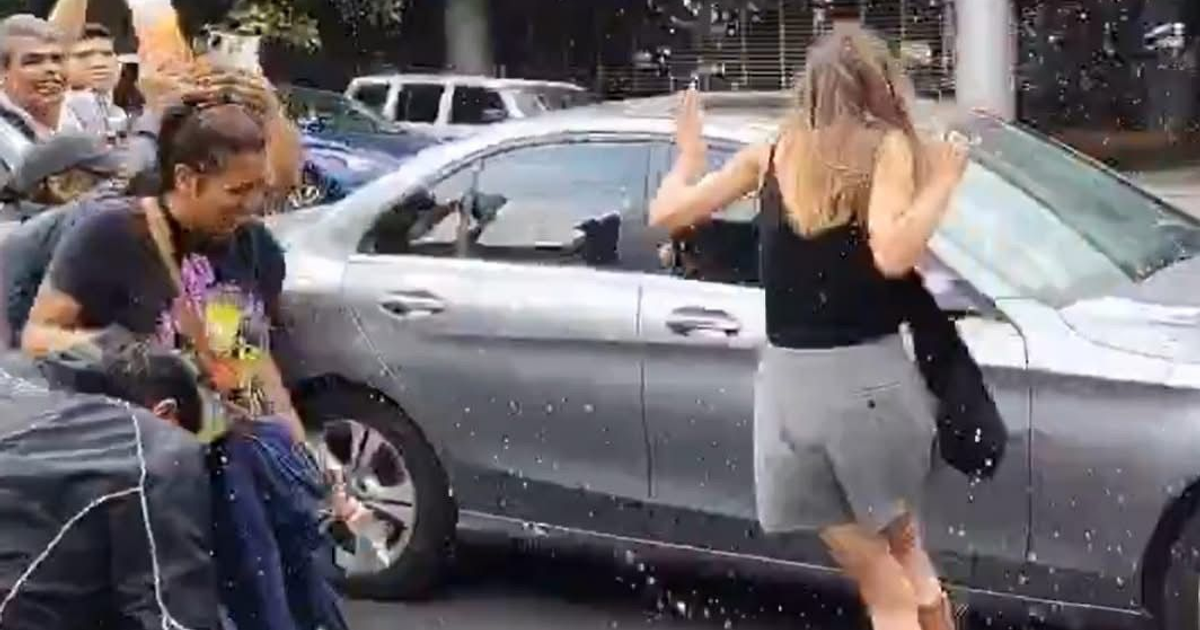 Personas interactuando cerca de un coche en una calle animada, con un ambiente festivo y un líquido derramado.