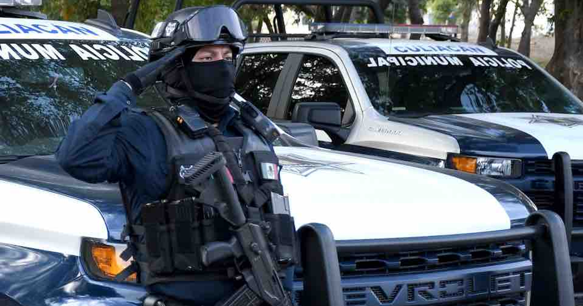 Oficial de policía con uniforme táctico saluda frente a vehículos de la Policía Municipal en Sinaloa.