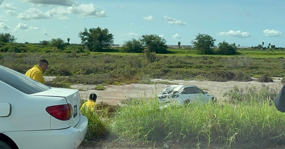 Dos hombres con camisetas amarillas junto a un vehículo blanco dañado en un accidente en carretera.