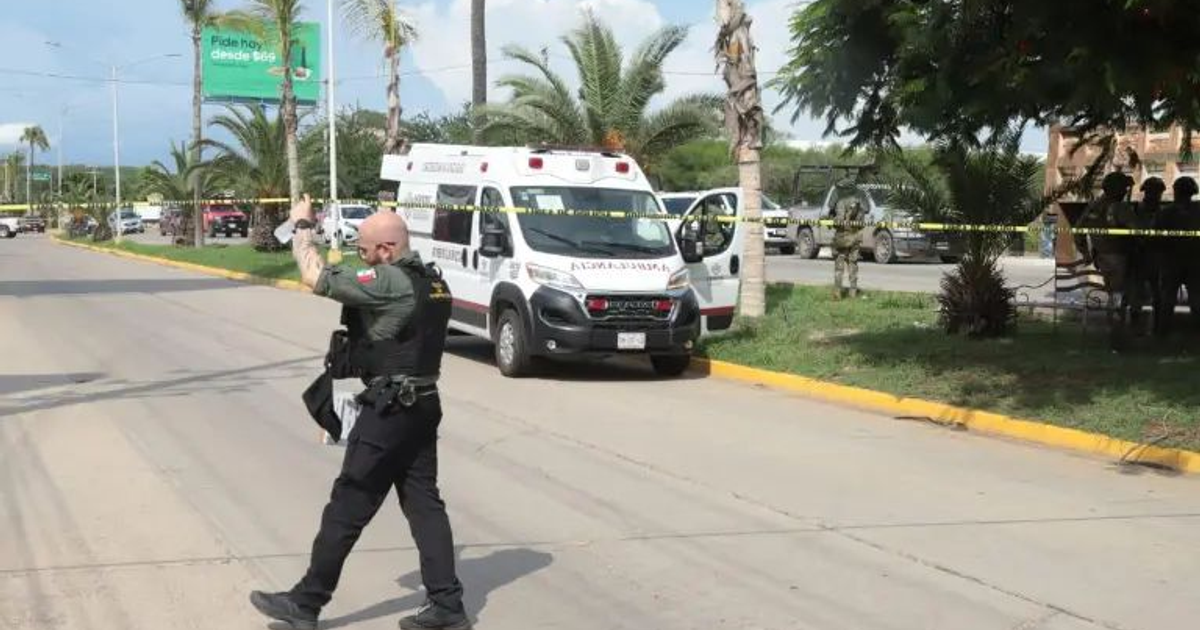 Oficial de policía asegurando la zona tras ataque armado a ambulancia en Mazatlán, con ambulancias y cinta amarilla en el fondo.