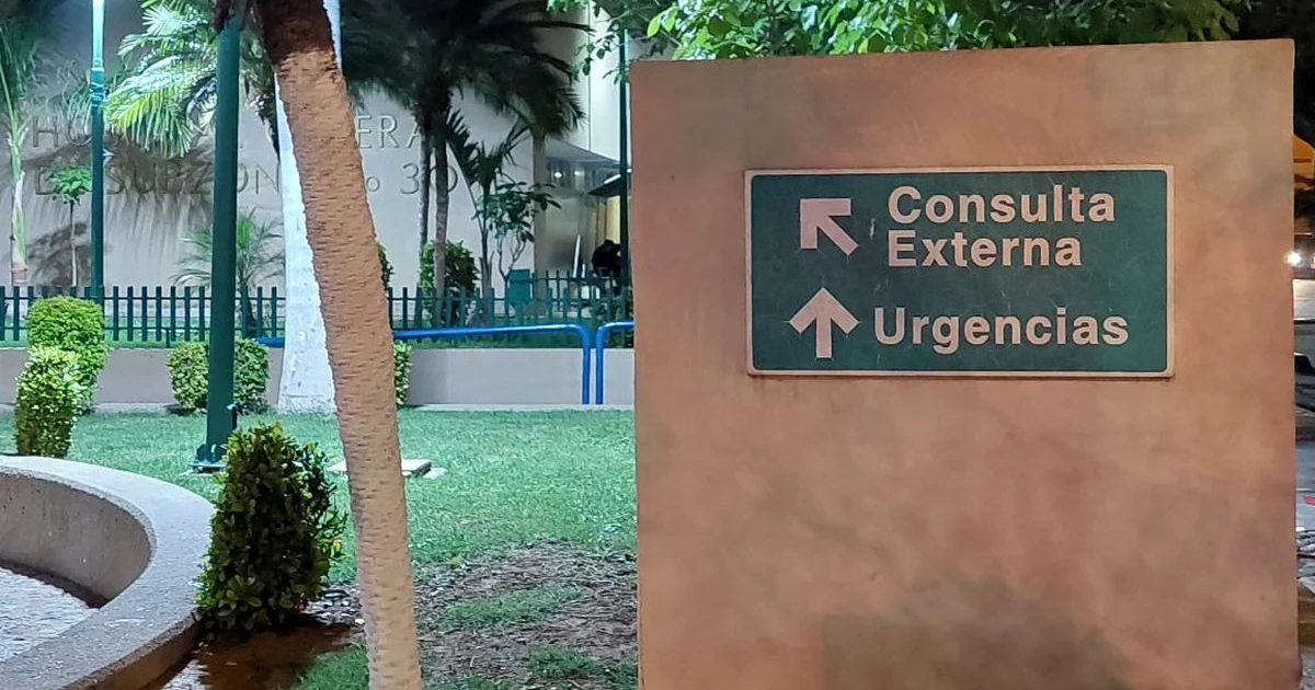 Letrero hospitalario indicando direcciones a Consulta Externa y Urgencias, con fondo de plantas y edificio.