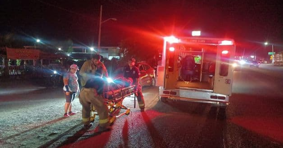 Paramédicos atienden a un adulto mayor herido en una escena nocturna de emergencia, con una ambulancia y luces intermitentes en el fondo.