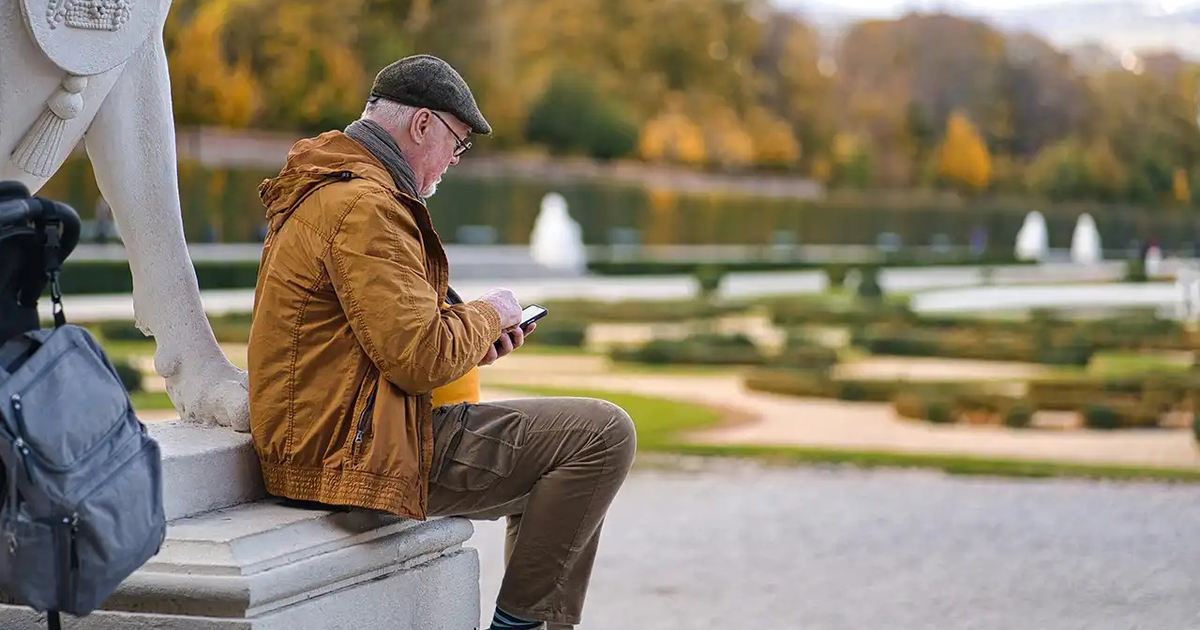 Hombre mayor con abrigo naranja sentado al aire libre mirando su teléfono, rodeado de un paisaje otoñal.