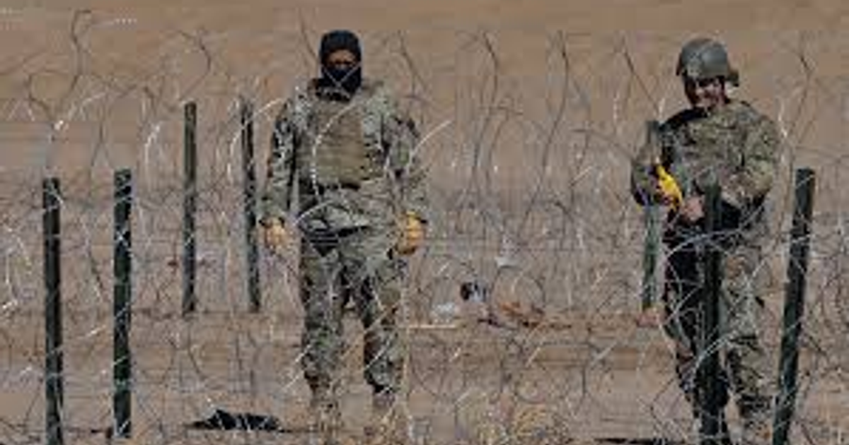 Dos soldados en un área árida con alambre de púas, uno sosteniendo un dispositivo, en un contexto de vigilancia fronteriza.