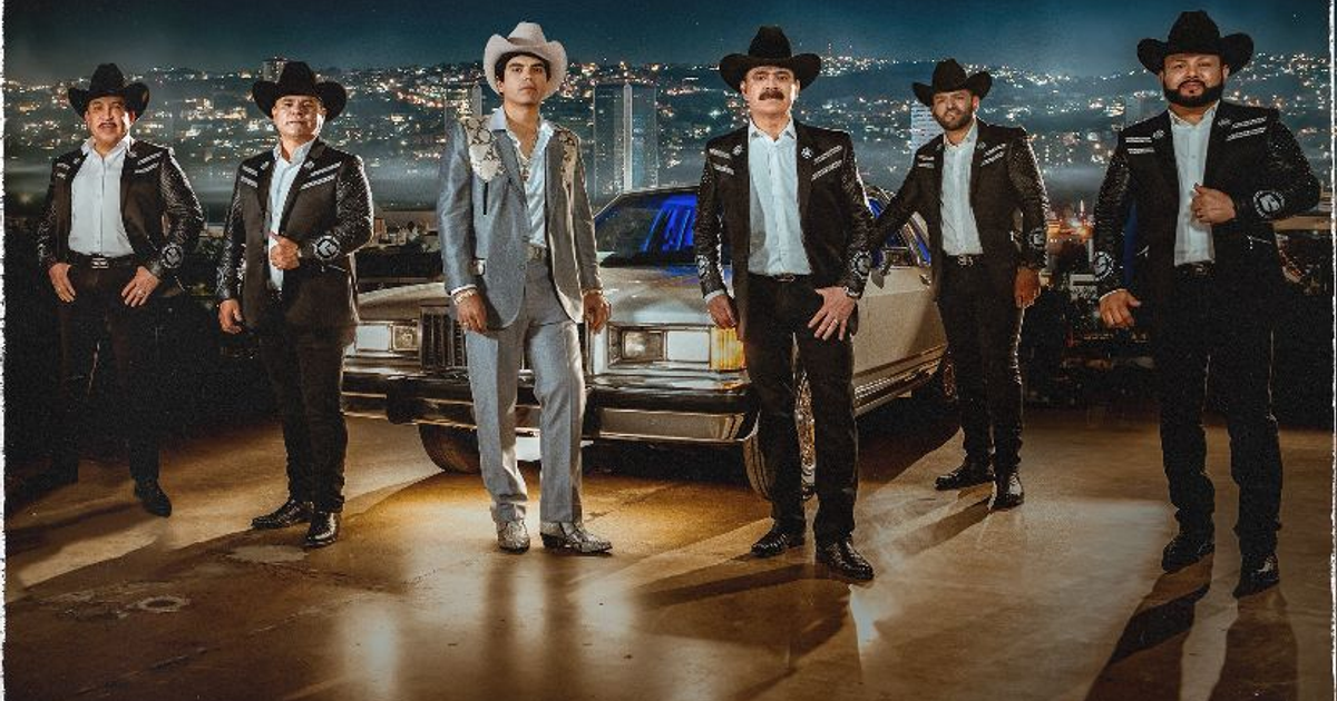 Grupo de músicos regionales mexicanos con trajes típicos y sombreros vaqueros posando frente a un automóvil clásico, con una ciudad iluminada al fondo.