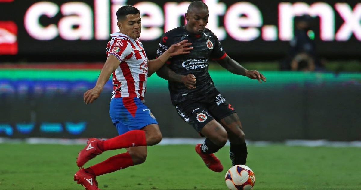 Dos jugadores de fútbol, uno con camiseta a rayas rojas y blancas y otro con uniforme negro, luchan por el balón en el campo durante el partido Chivas vs Xolos.