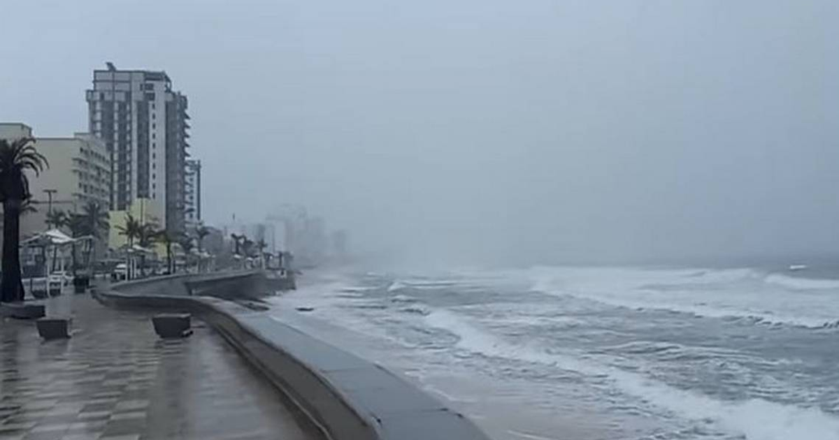 Paisaje costero en Sinaloa con clima tormentoso, edificios altos, palmeras y olas rompiendo en la orilla.