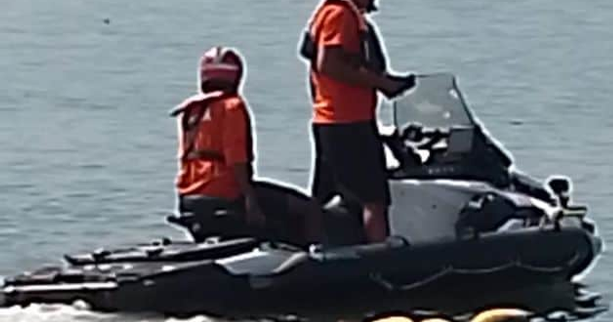 Dos personas en moto de agua, una de pie al mando y otra sentada, ambas con ropa naranja, en un entorno acuático.