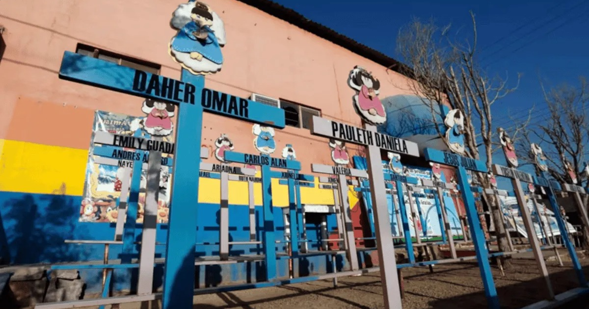 Muro rosa con cruces y figuras humanas, simbolizando comunidad y memoria, con cielo despejado y árboles invernales de fondo.