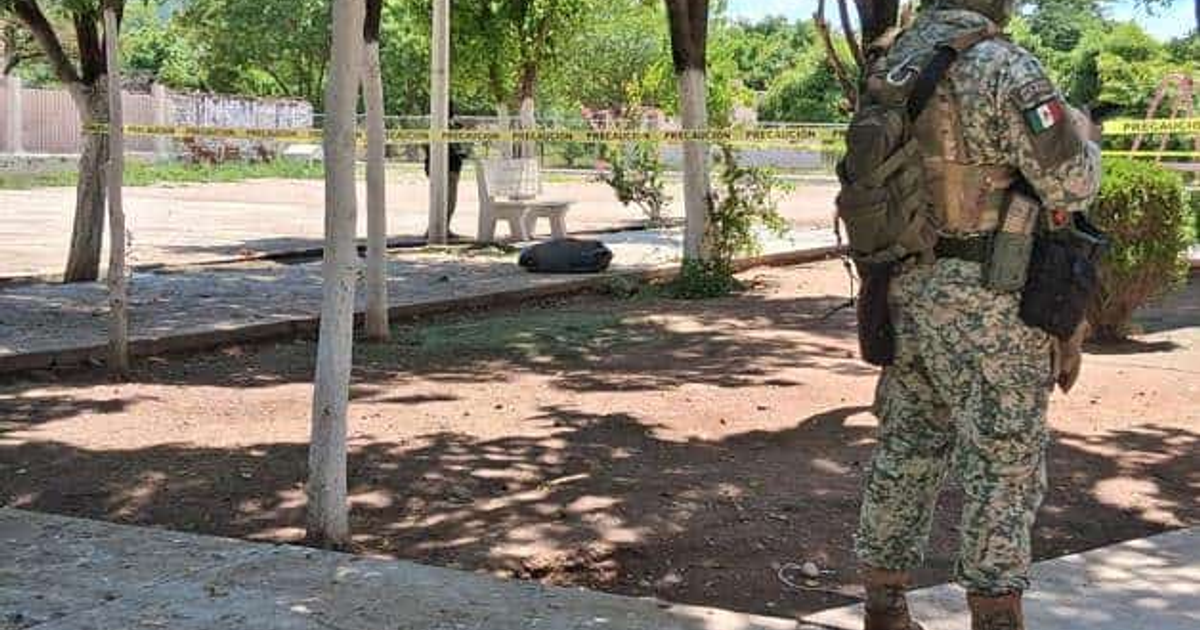 Soldado en uniforme camuflado en un área acordonada con cinta de precaución, rodeado de árboles y un banco de concreto.