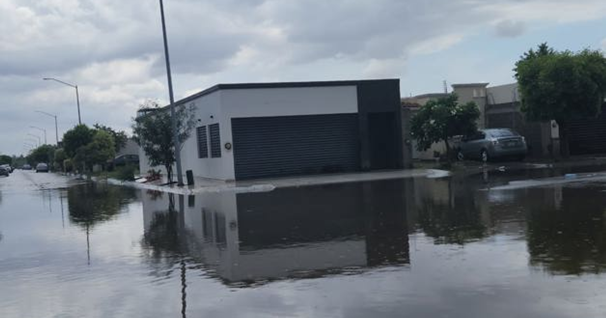 Calle inundada en Los Mochis con reflejo de edificios y árboles, automóvil aparcado y cielo nublado.