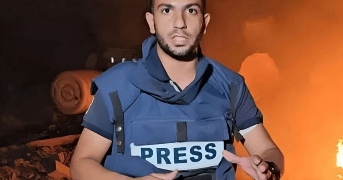 Periodista con chaleco de prensa reportando en zona de conflicto afectada por fuego o humo.