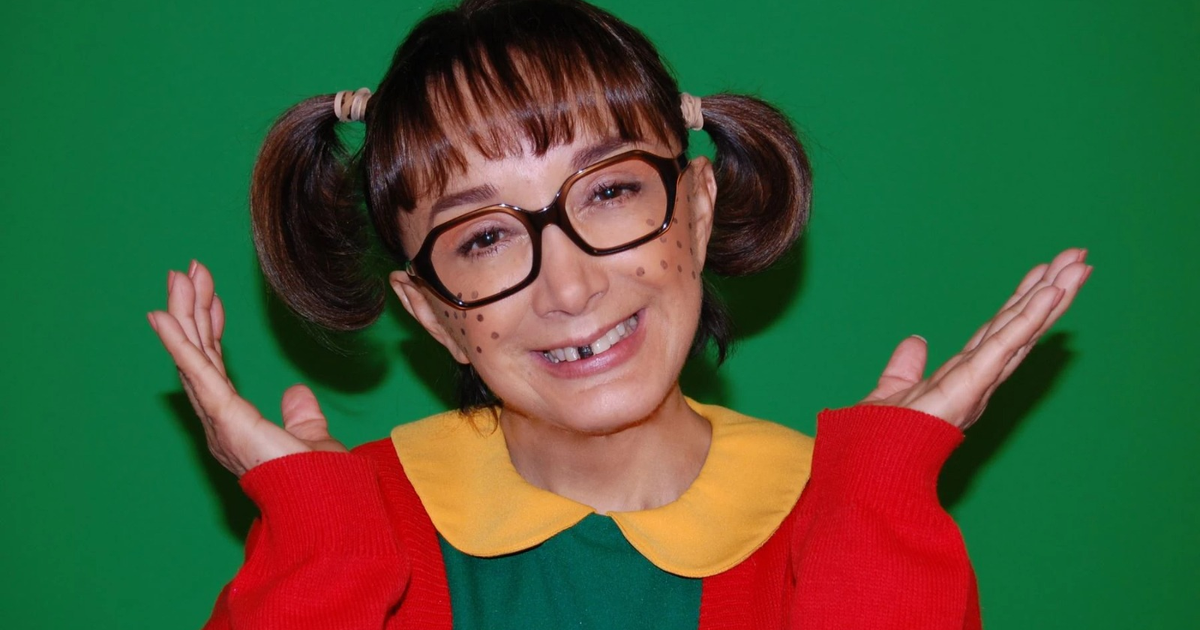 Persona disfrazada de La Chilindrina con gafas grandes, camiseta verde, suéter rojo y coletas, sonriendo sobre fondo verde.