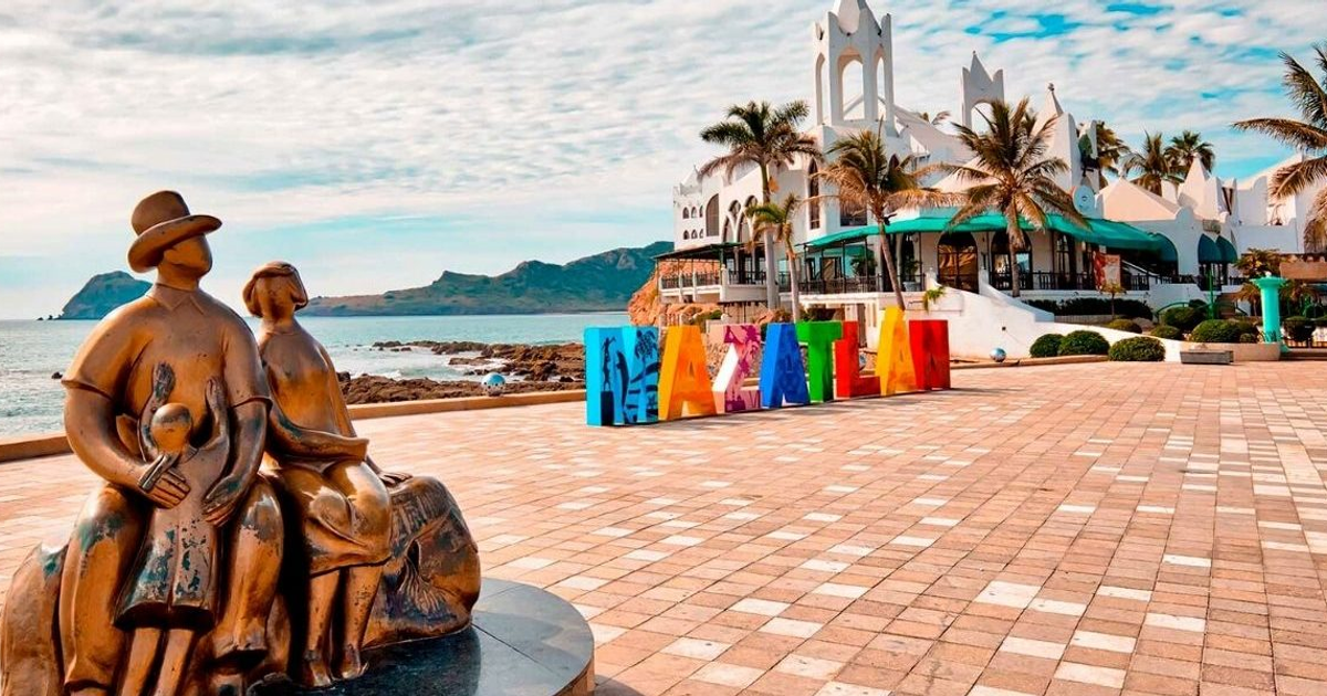Escultura familiar de bronce en Mazatlán con mar y montañas al fondo, edificio blanco a la derecha y letrero 'MAZATLÁN'.