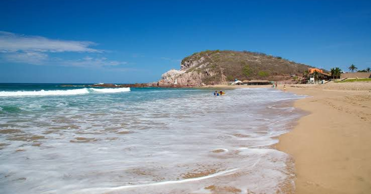 Playa de Mazatlán con arena dorada, aguas claras y colina verde al fondo, cerca del hotel Park Inn.