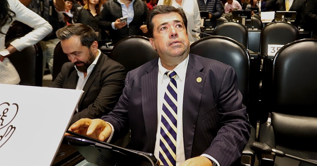 Dos hombres en un foro político, uno usando un dispositivo electrónico, con una audiencia al fondo.