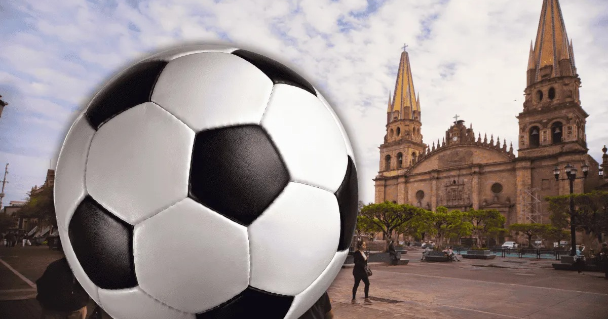 Balón de fútbol en primer plano con iglesia y cielo nublado al fondo, personas en la plaza.