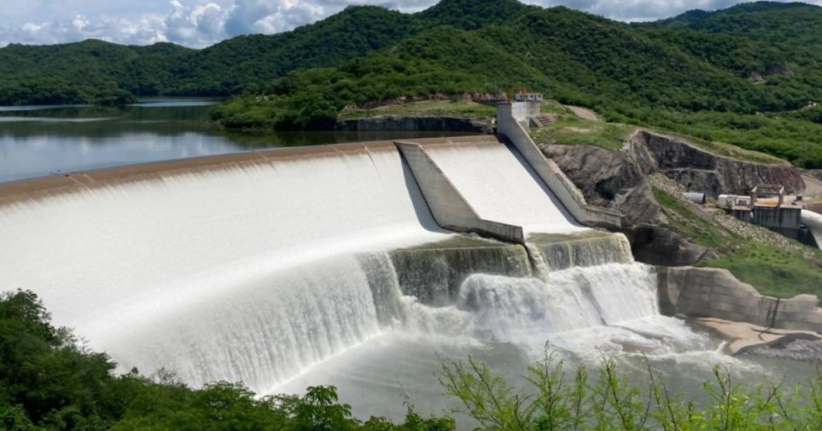 Vista de la Presa Picachos con agua cayendo por el vertedero, colinas y vegetación al fondo.