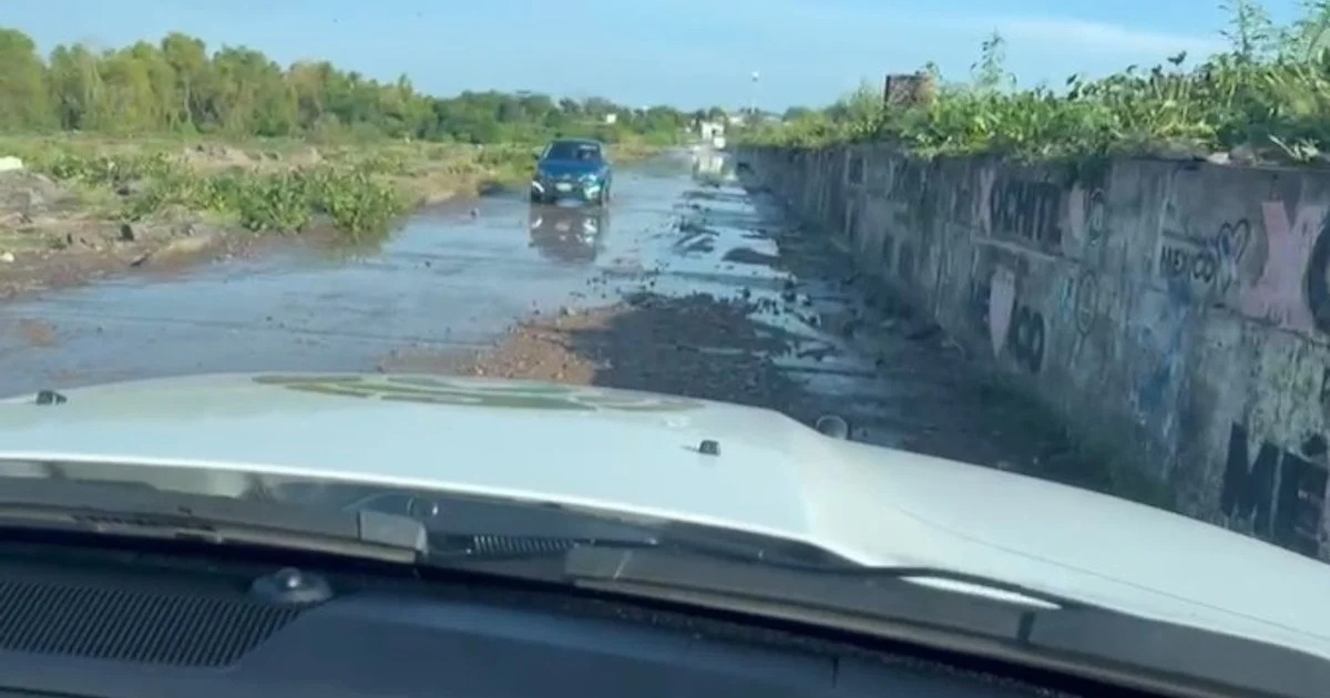 Carretera con agua estancada y barro en Culiacán, automóvil visible, vegetación y muro con grafitis, cielo despejado.